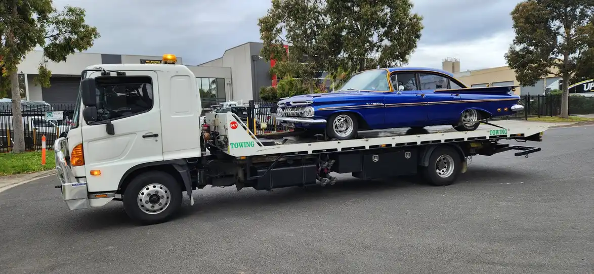 A white tilty tray tow truck transporting a blue vintage car on an urban street, with commercial buildings and trees in the background.