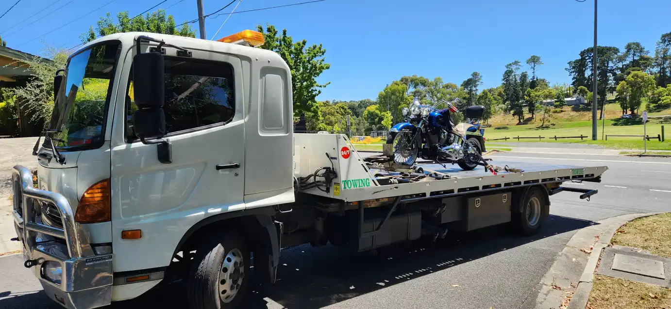 A white tilty tray tow truck transporting a blue cruiser motorcycle on a sunny day, parked near a grassy area with trees and clear blue skies.