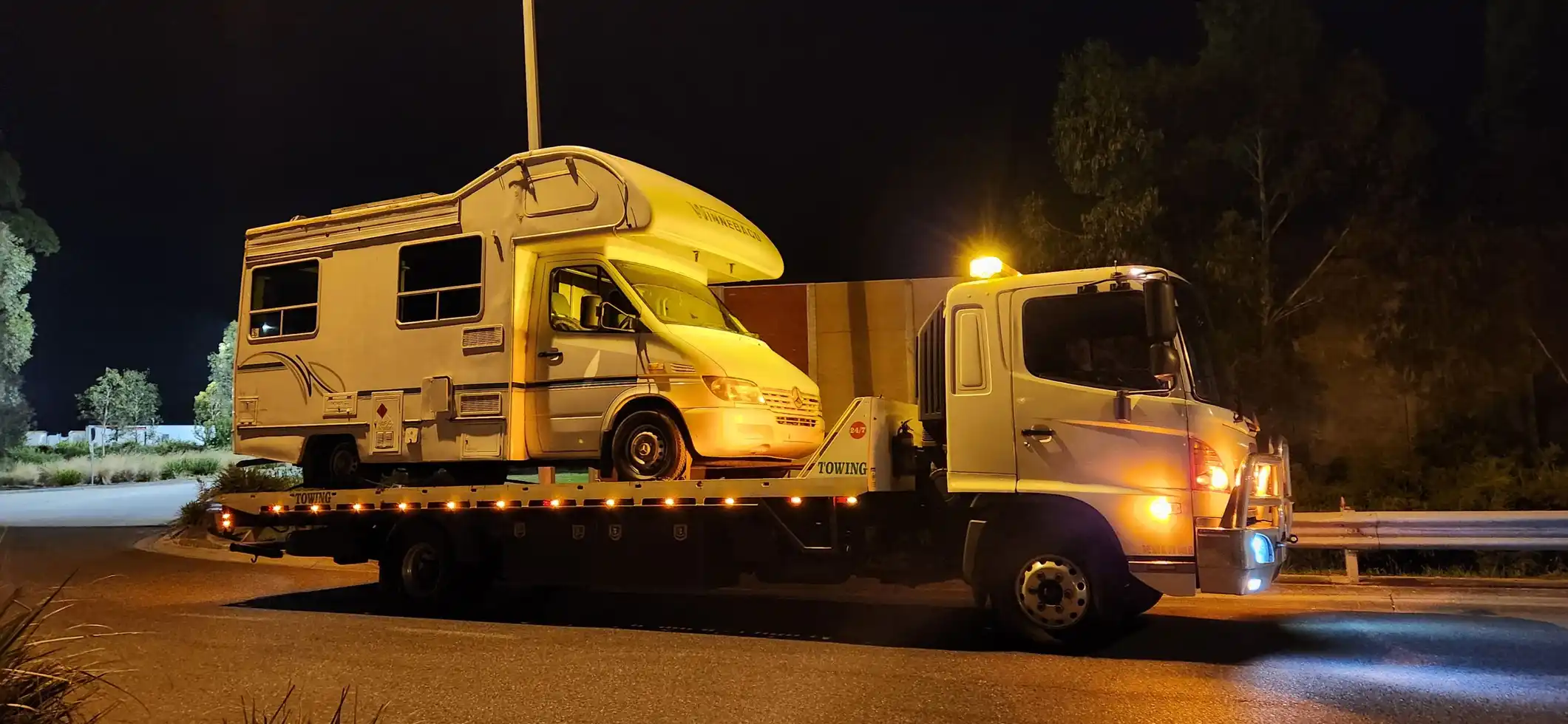 A tilty tray tow truck carrying a large white motorhome under bright lights at night, parked beside a road with trees and a dark sky in the background.