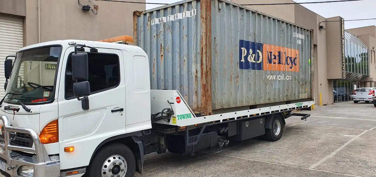 A white flatbed tow truck loaded with a large, weathered shipping container in an industrial area with office buildings in the background.